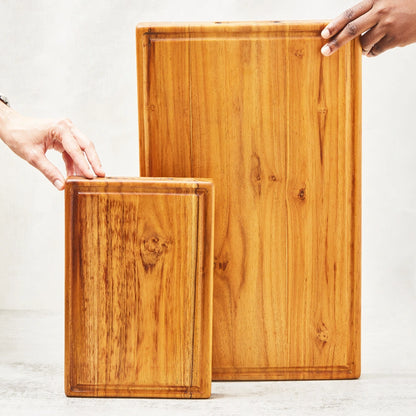 One large and one small premium teak cutting board with a juice groove and embedded handle, standing upright to display their main surfaces, made from 100% certified teak wood.